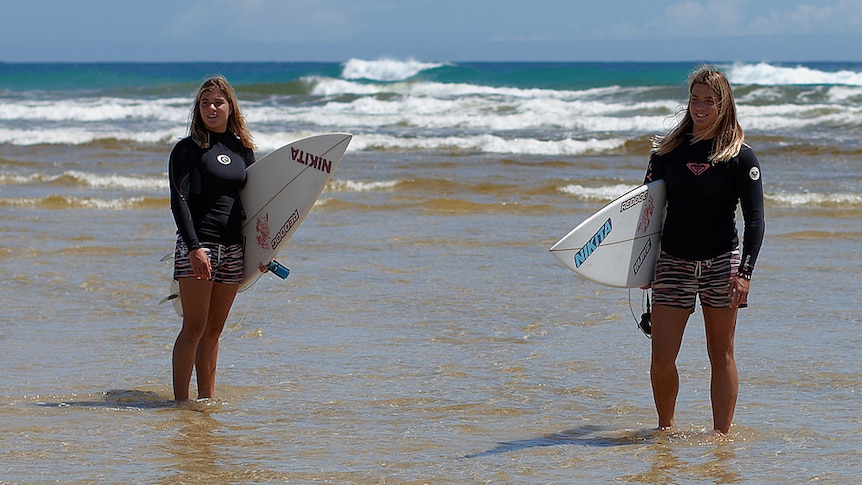 A pair of identical twins stand in the shallows on a beach, holding surfboards.