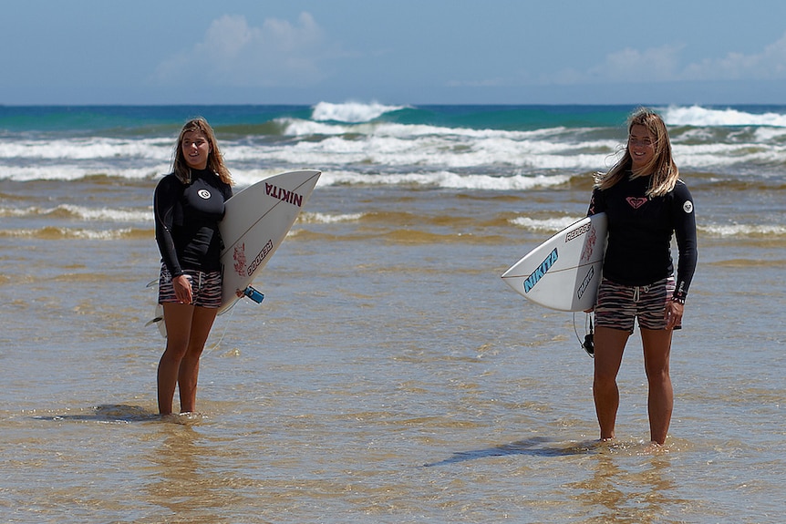 A pair of identical twins stand in the shallows on a beach, holding surfboards.