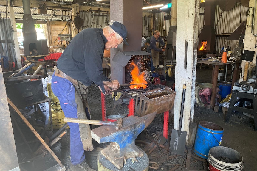 An elderly man stands over a blacksmith forge with flames