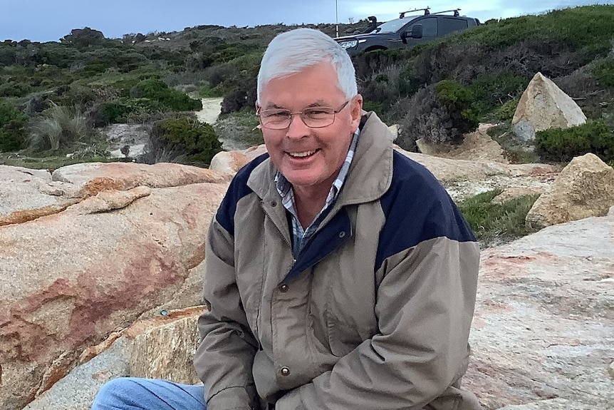 A smiling, grey-haired man sits on a beach beneath a stormy sky.