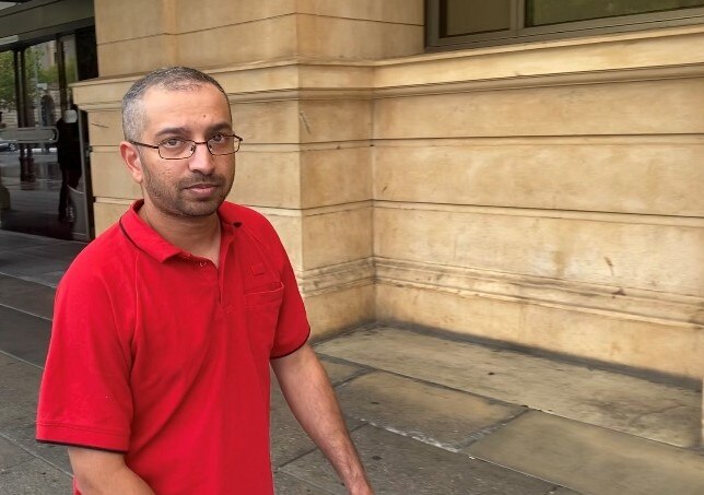 A man in a red t-shirt and glasses looks directly at the camera while walking outside a court building