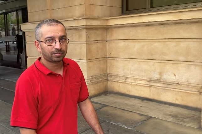 A man in a red t-shirt and glasses looks directly at the camera while walking outside a court building
