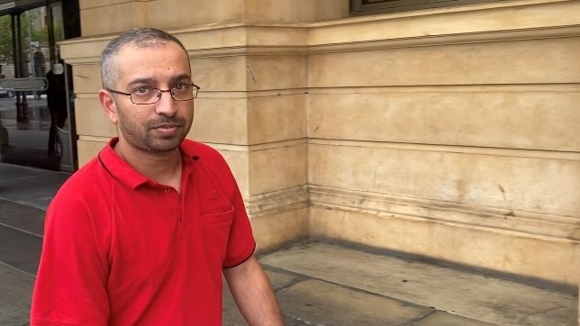 A man in a red t-shirt and glasses looks directly at the camera while walking outside a court building