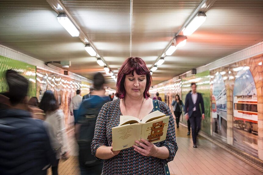 Colour photograph of woman standing still reading a book, people pass her by in a blur.