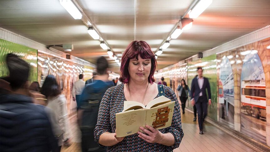 Colour photograph of woman standing still reading a book, people pass her by in a blur.