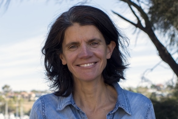 A woman with shoulder-length dark hair and a denim shirt stands smiling at the camera