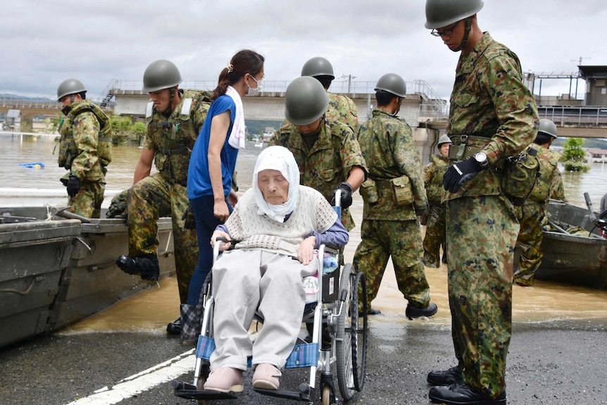 Military personnel push an elderly woman in a wheelchair with flooding in the background.