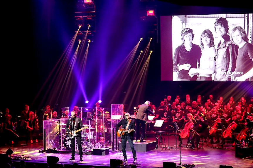 A rock band plays on stage in front of a symphony orchestra