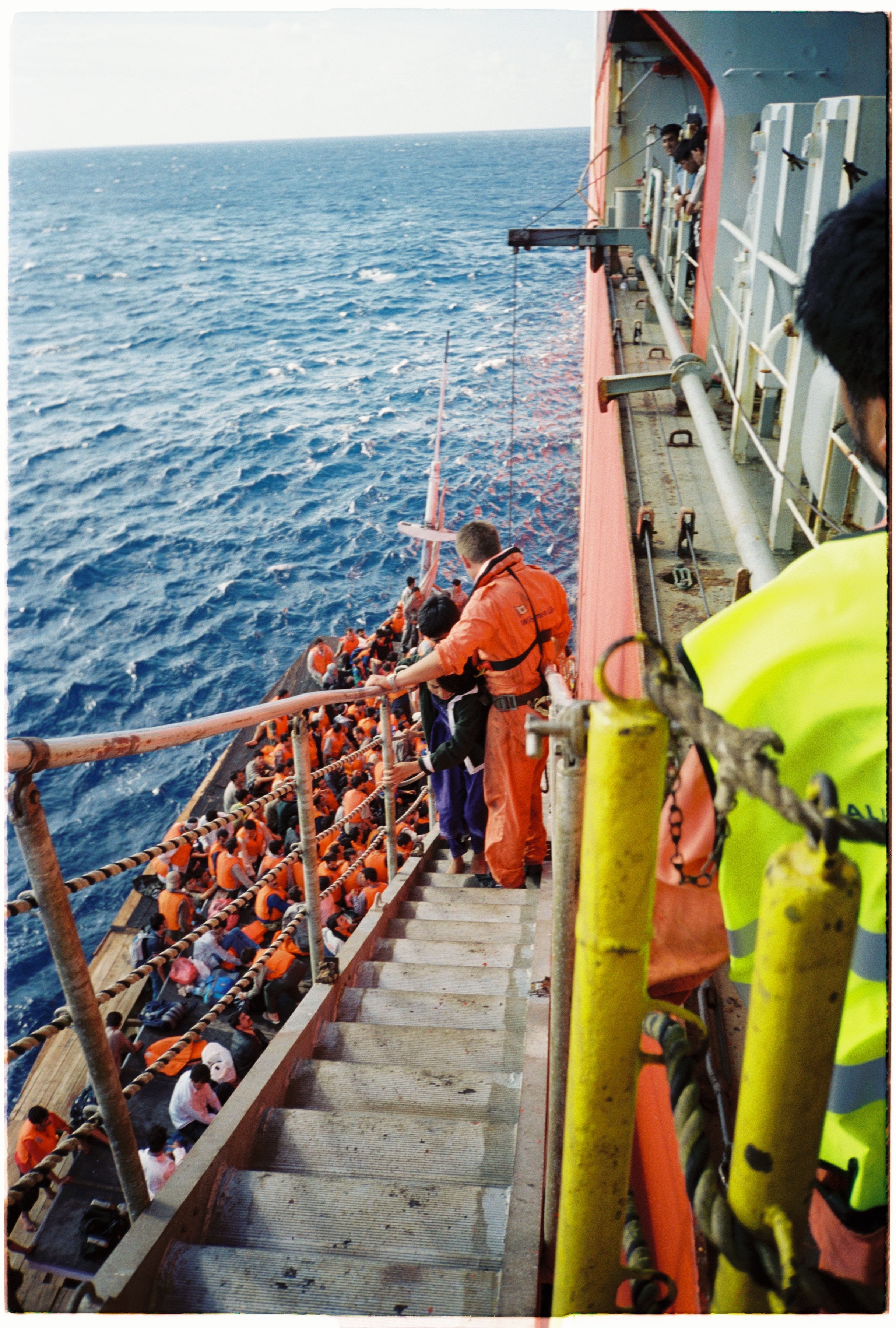 People in red life jackets climb from a small wooden boat up some stairs to the side of a freighter.