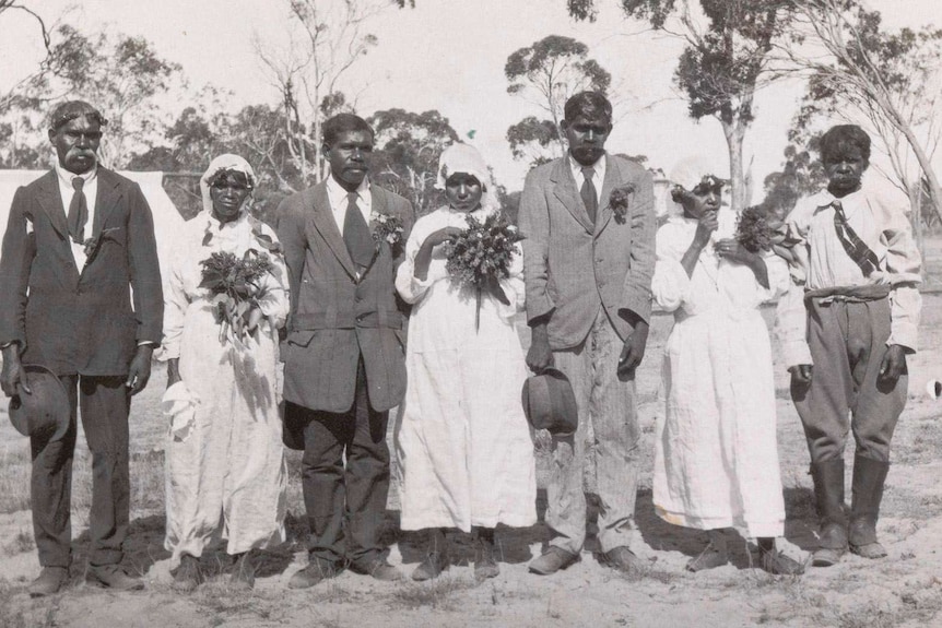 four aboriginal men in suits and three aboriginal women in white wedding dresses