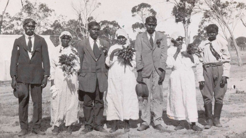 four aboriginal men in suits and three aboriginal women in white wedding dresses