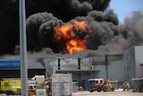 Thick black smoke and flames rise into the sky above an industrial waste facility with fire trucks and crews below.