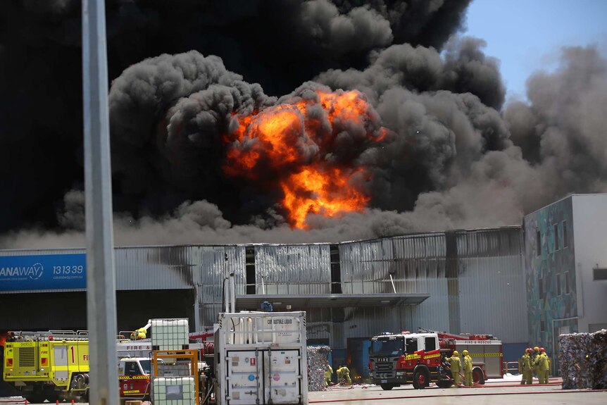 Thick black smoke and flames rise into the sky above an industrial waste facility with fire trucks and crews below.