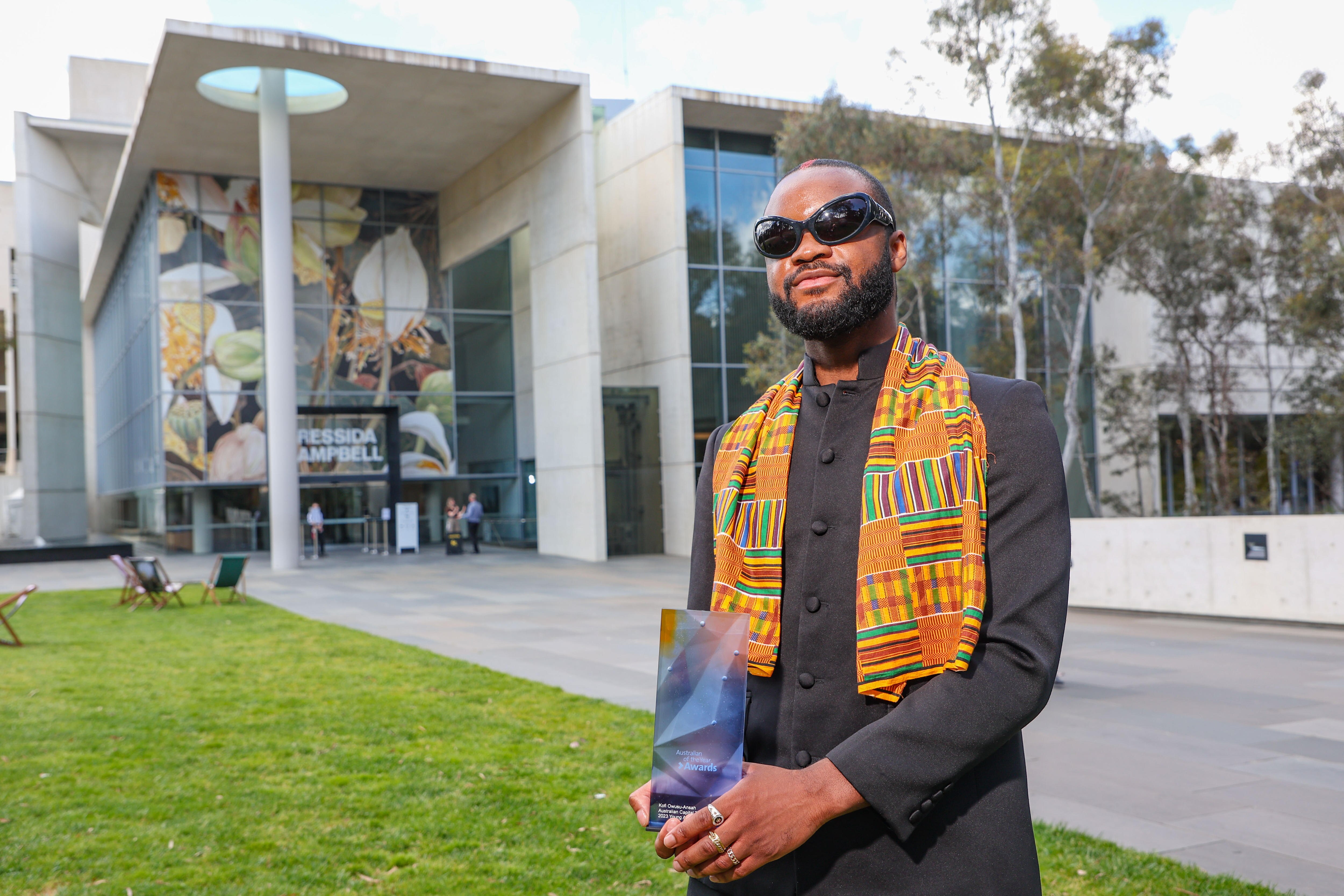 Man in big black glasses and colourful, patterned scarf holding his australian of the year award 