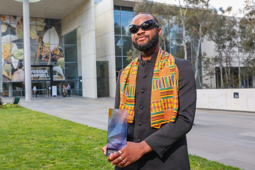 Man in big black glasses and colourful, patterned scarf holding his australian of the year award 