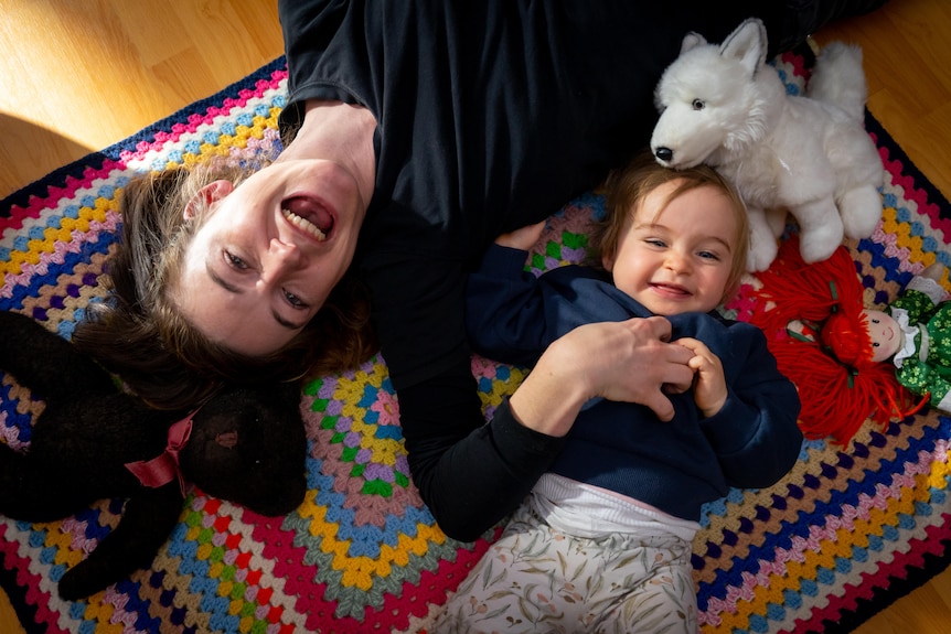 A woman and a baby laughing and lying on a rug next to toys