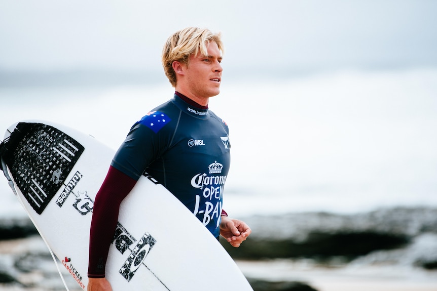 Ethan Ewing, wearing a dark wetsuit, walks while holding a white surf board.