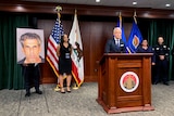 A man at podium, flanked by three other people and various flags, stands beside a photo of a man charged with sexual assault