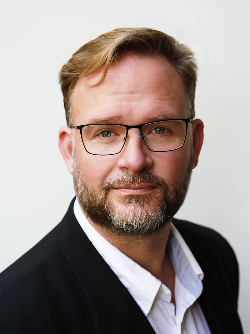 Professional studio portrait of a man in an open-collared shirt with glasses.