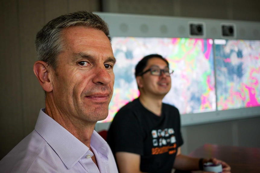 Man in purple business shirt looks into camera while sitting at round meeting table, another man in background