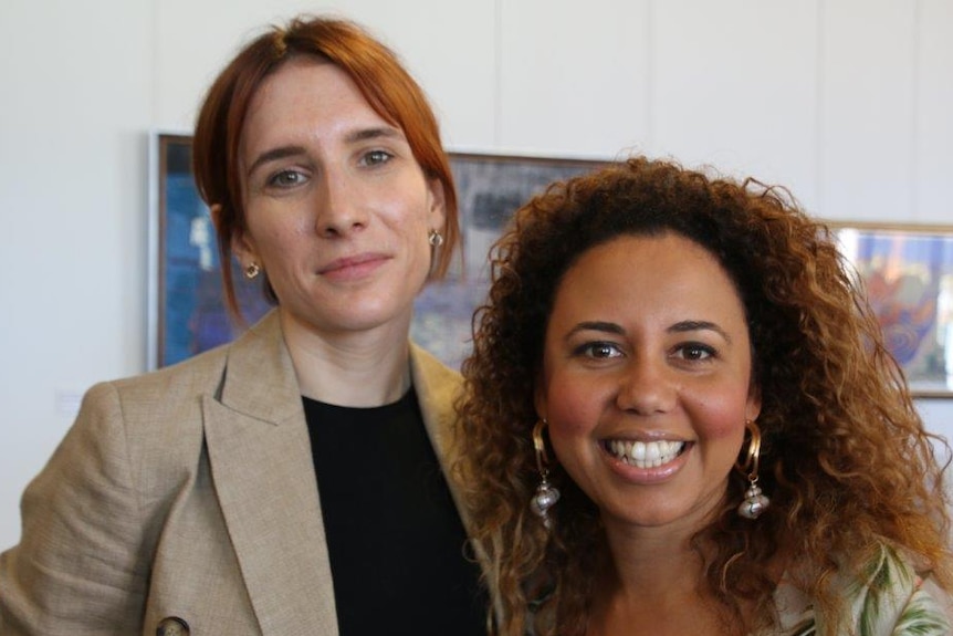 A close up of Martina and Jessica standing close together in a white room, looking to camera, with paintings on the wall behind.