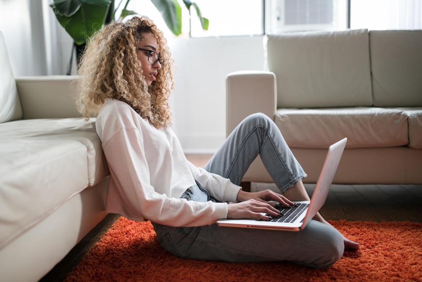 A young woman with curly hair sits on the ground propped up against a couch with a laptop on her lap. 