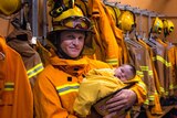 A firefighter decked out in full uniform poses for a photo while cradling his sleeping newborn son
