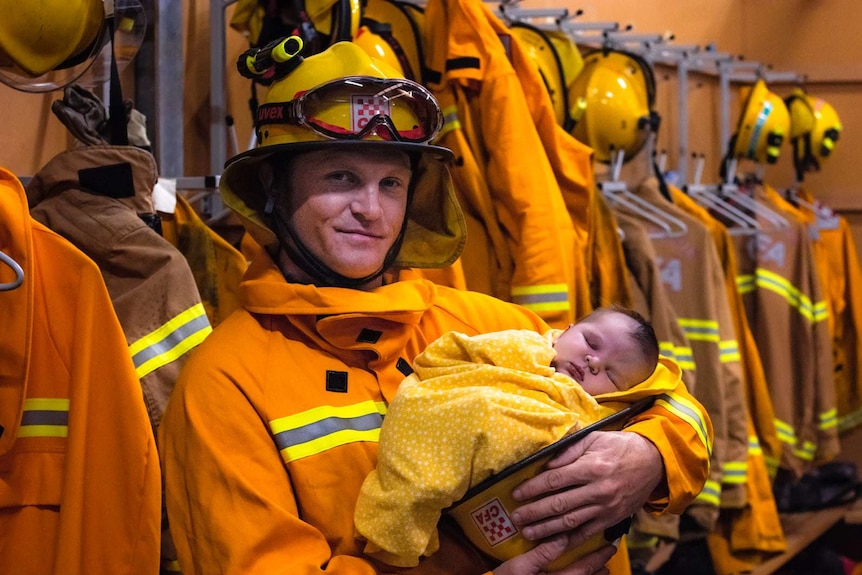A firefighter decked out in full uniform poses for a photo while cradling his sleeping newborn son