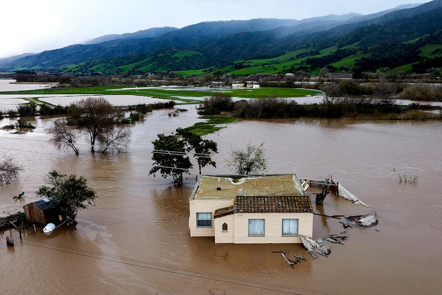 floodwater surrounds a house near mountaings in california, with trees poking out of the water