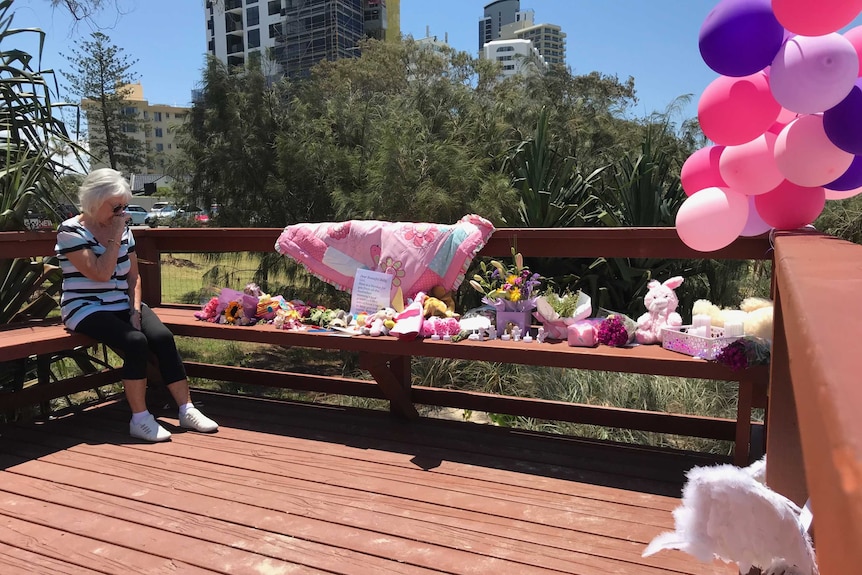 Woman sits emotionally, looking at the memorial of flowers and toys.