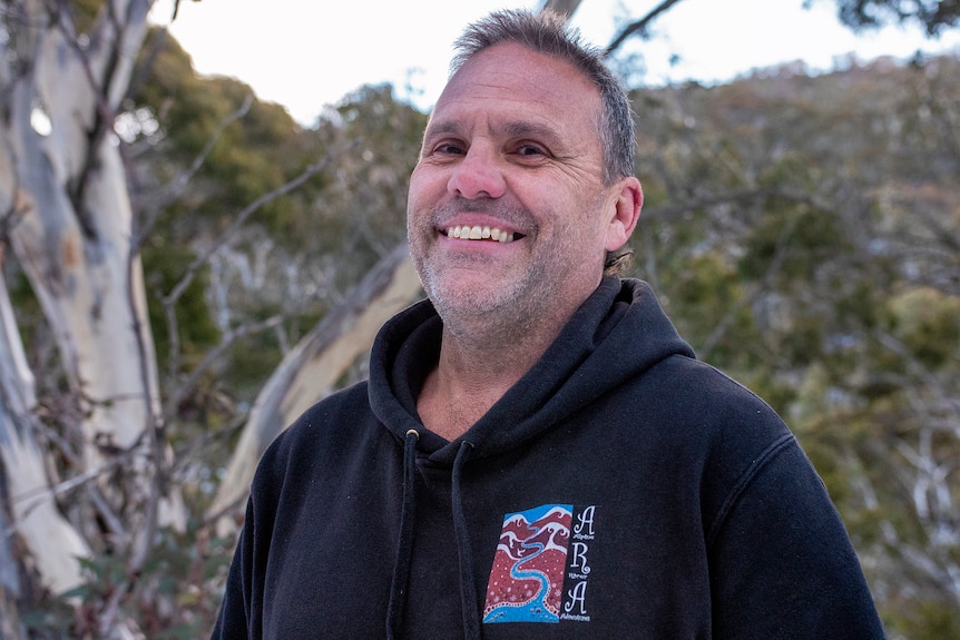 A man wearing a black hoodie, short grey hair stands among gum trees smiling.