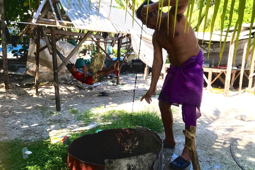 A man pulls a string out of a well in a wood and corrugated iron structure with a person lying in a hammock in the background.