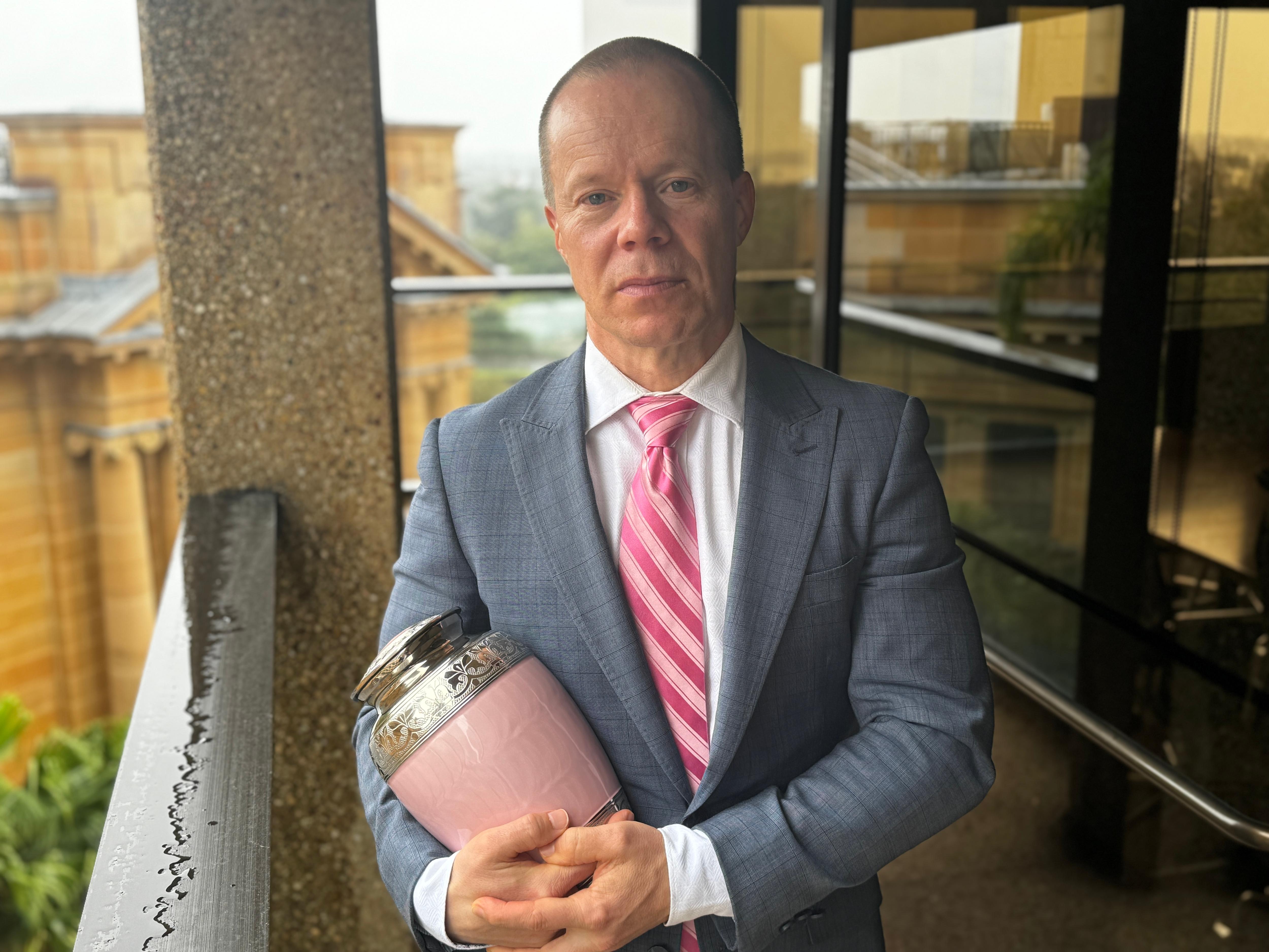 A middle-aged man in a suit standing on a balcony looks out towards the sky while holding a pink urn.