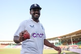 England bowler Rehan Ahmed holds a cricket ball and smiles for the camera as he walks off the field during a Test match.