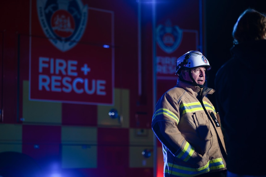 A man in protective fire gear stands in front of a fire and rescue truck