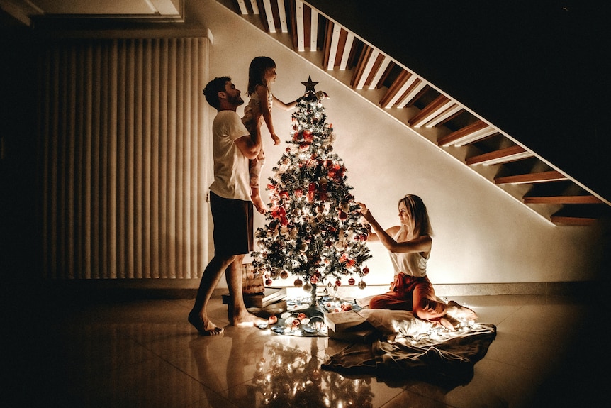 一个男人、一个女人和一个孩子正在装饰一棵只有仙女灯点亮的塑料圣诞树。  