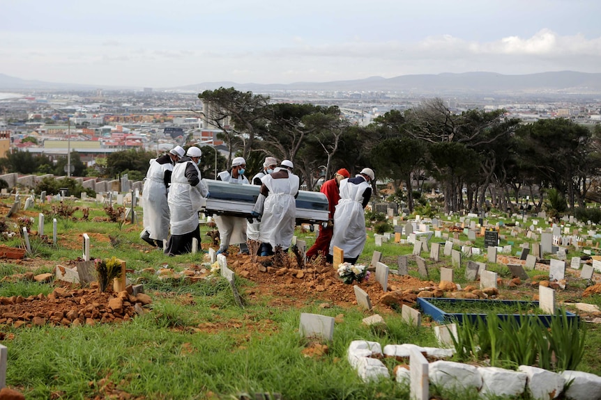 A group of men in PPE carry a coffin through a graveyard