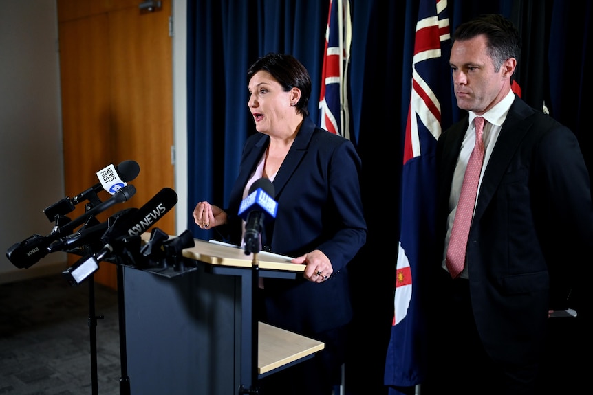 a man standing behind a woman who is talking at a podium 