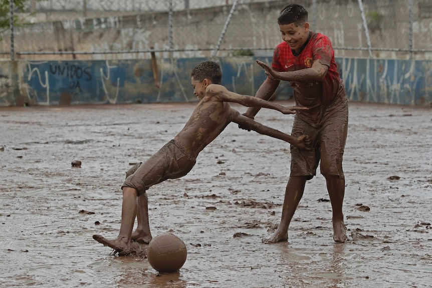 On a brown, mud-soaked street, two boys play soccer with one of them slipping on the ground.