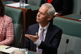 Giles gestures with one hand and looks confused, sitting in the lower house.