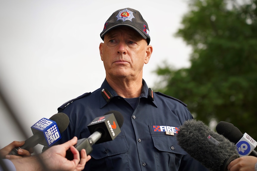 A man wearing a navy MFS uniform and cap stands in front of media microphones