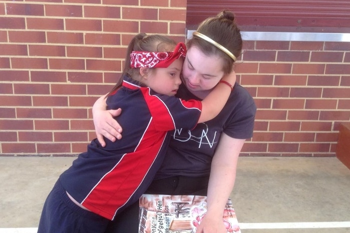 Child in uniform with bandanna hugs lady sitting in a chair holding a book.