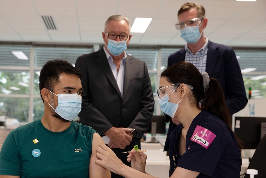 a man gets a vaccine while two other men watch on