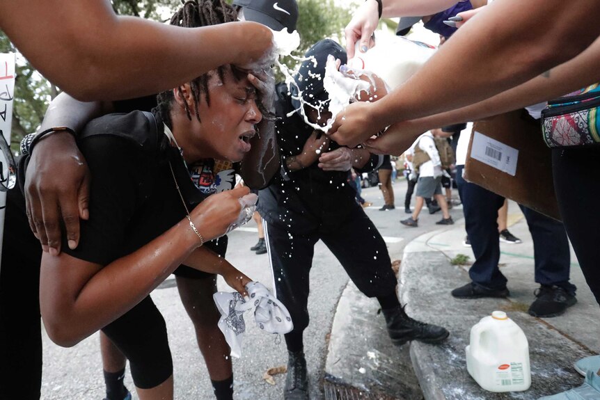 A woman stands bending over as people help her by pouring milk onto her face to offset tear gas effects.