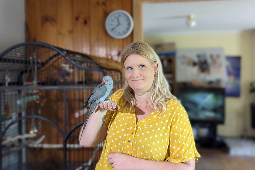 Shelley, a woman with blonde hair and a yellow top, smiles as a pet bird sits on her hand.