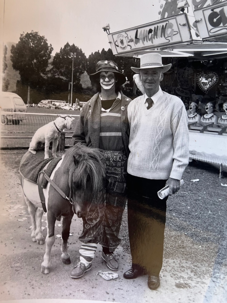 A woman dressed as a clown next to a man in a QLD hat and a dog atop a pony