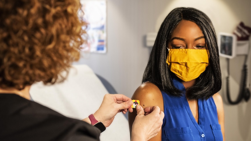 A woman wearing a bright yellow face mask look down at a nurse putting a sticker bandaid on her arm after a vaccination.