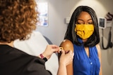 A woman wearing a bright yellow face mask look down at a nurse putting a sticker bandaid on her arm after a vaccination.
