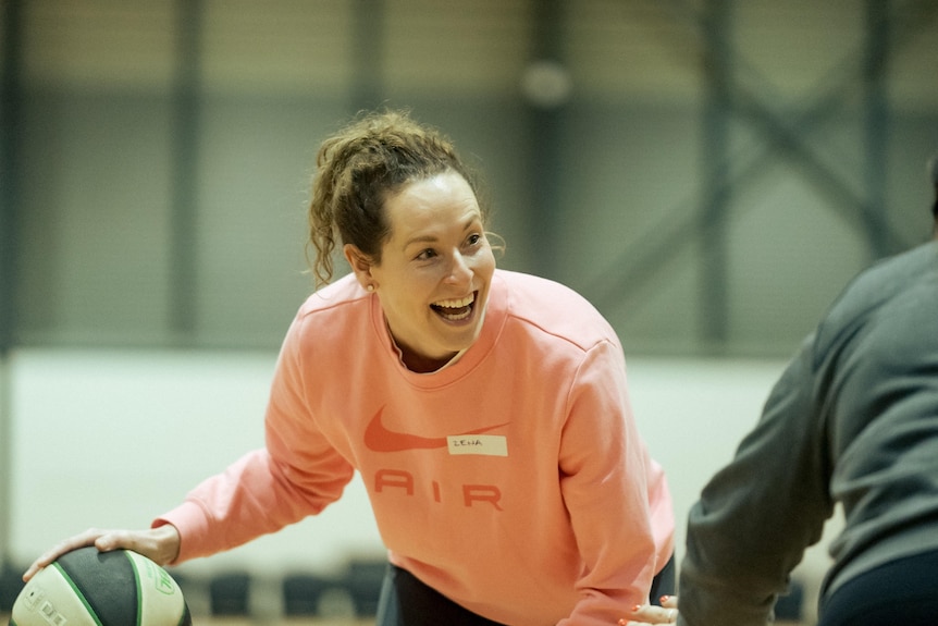 A woman wearing a pink jumper is smiling as she bounces a basketball.
