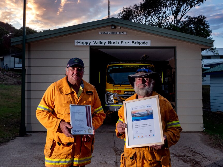 two men stand in yellow firefighting uniforms in front of the happy valley fire brigade